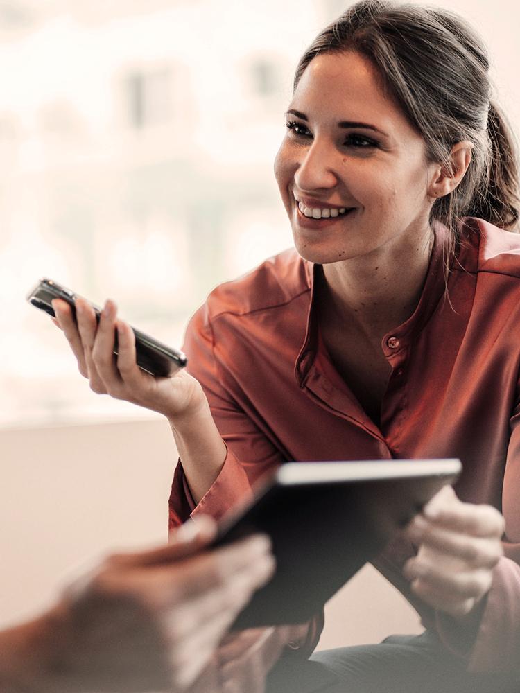 Mitarbeiterin mit Pferdeschwanz und rote Bluse hält Mobiles Telefon und lächelt Kollegen an.